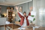 Shot of a young man celebrating while going over paperwork and using a laptop at home.