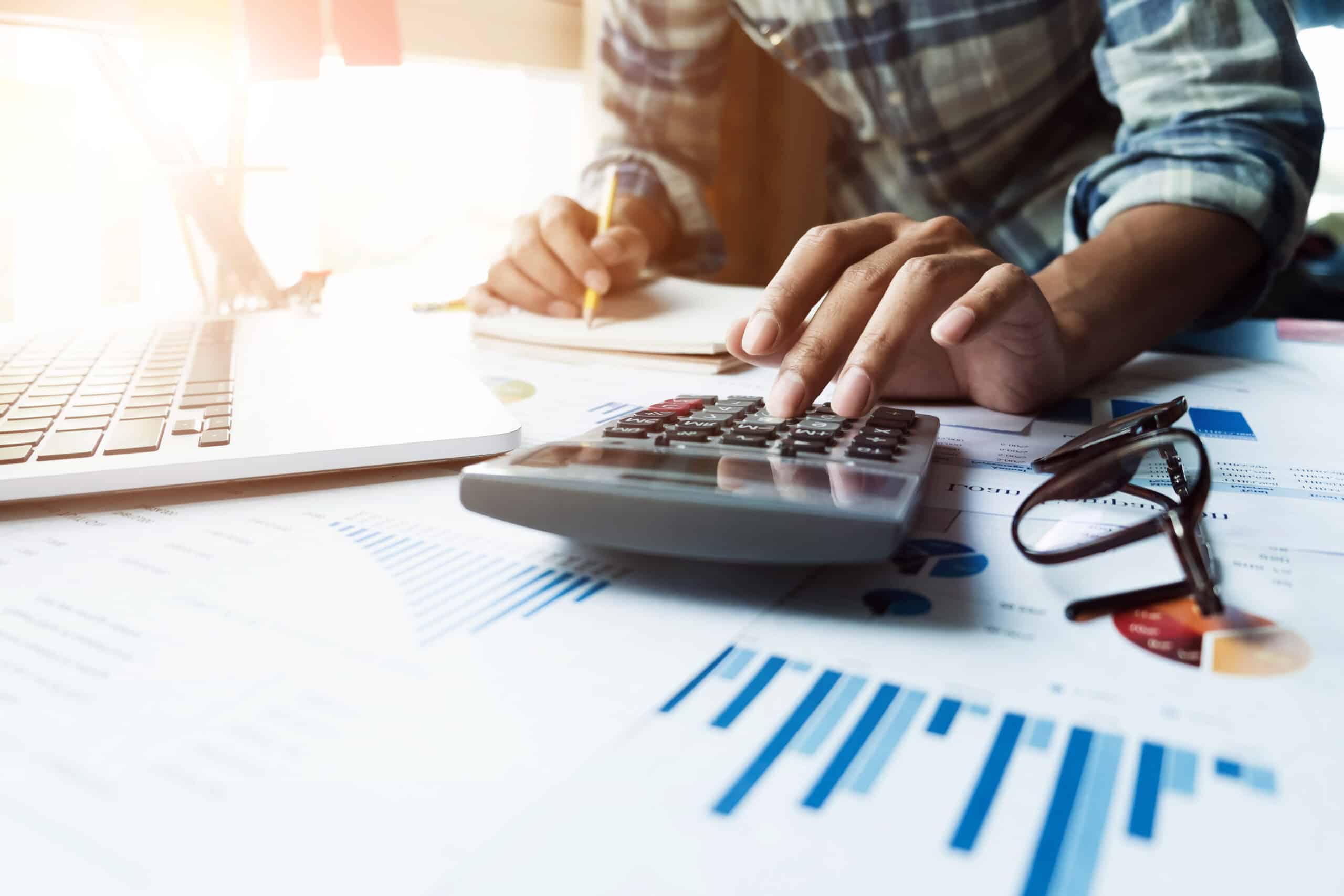 Close up of businessman or accountant hand holding pen working on calculator and laptop computer.