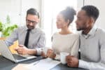 A tax professional sits and explains his findings to a young couple in his office in front of a laptop.