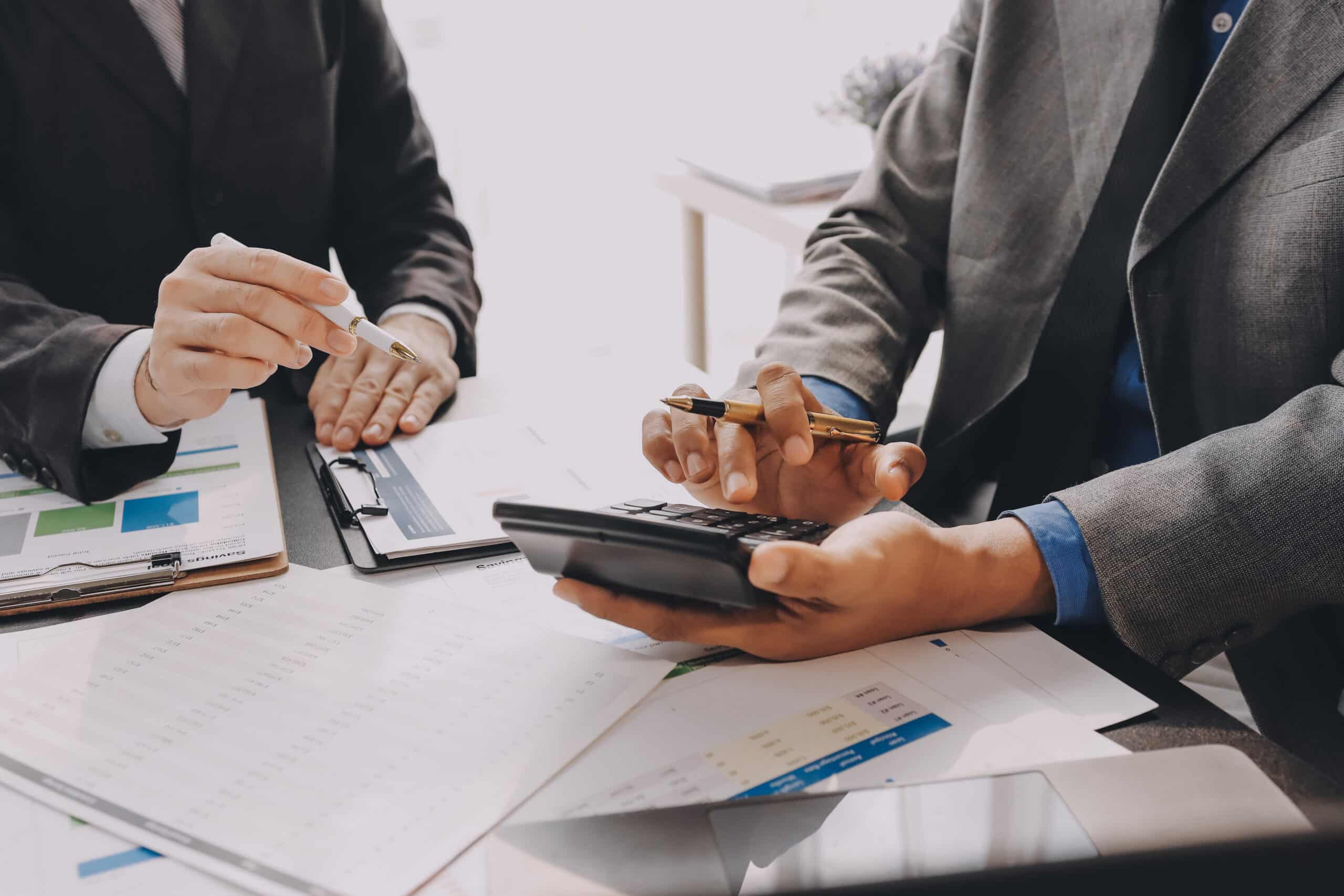 Two tax workers sit at a desk with binders, paperwork, and a calculator.