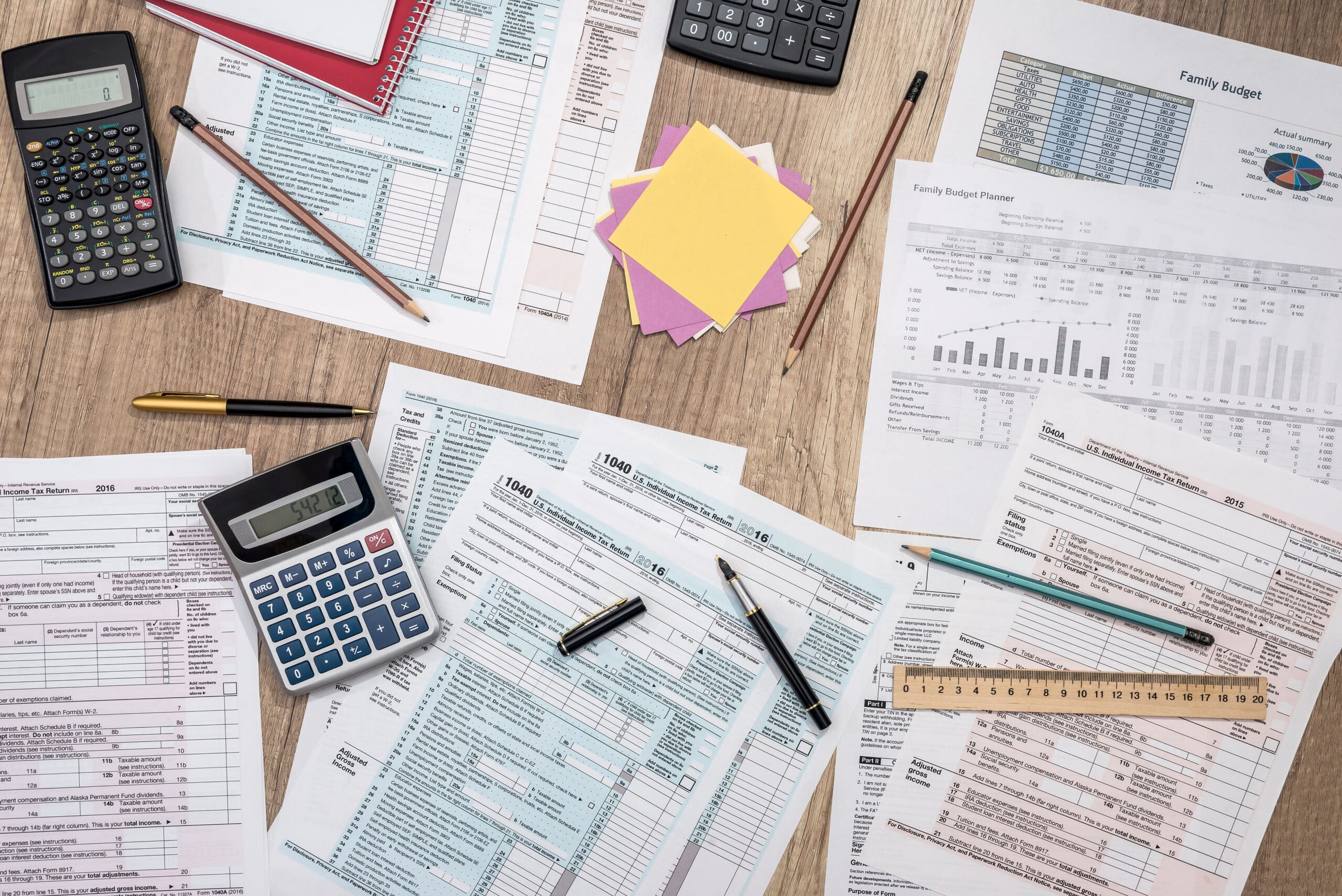 Assorted financial and tax paperwork sits scattered on a wooden desk with pens and calculators.