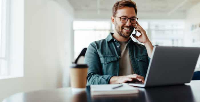 A man sits at a desk with coffee, a laptop, and a notebook while speaking happily on the phone.